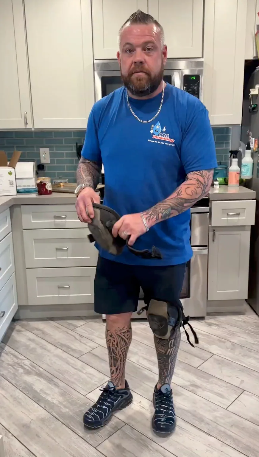 An MC Plumbing crew member holding knee pads in a kitchen, wearing a blue company T-shirt.