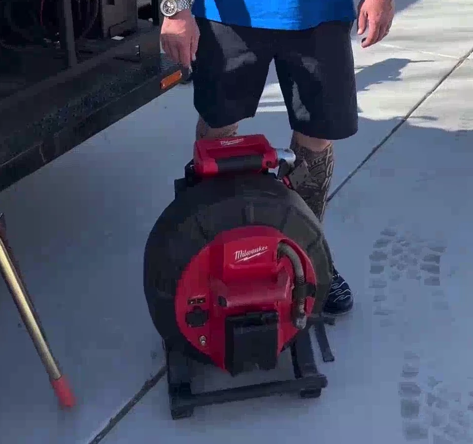 A red drain-and-sewer equipment reel on the concrete beside MC Plumbing's trailer, with a crew member standing behind it.