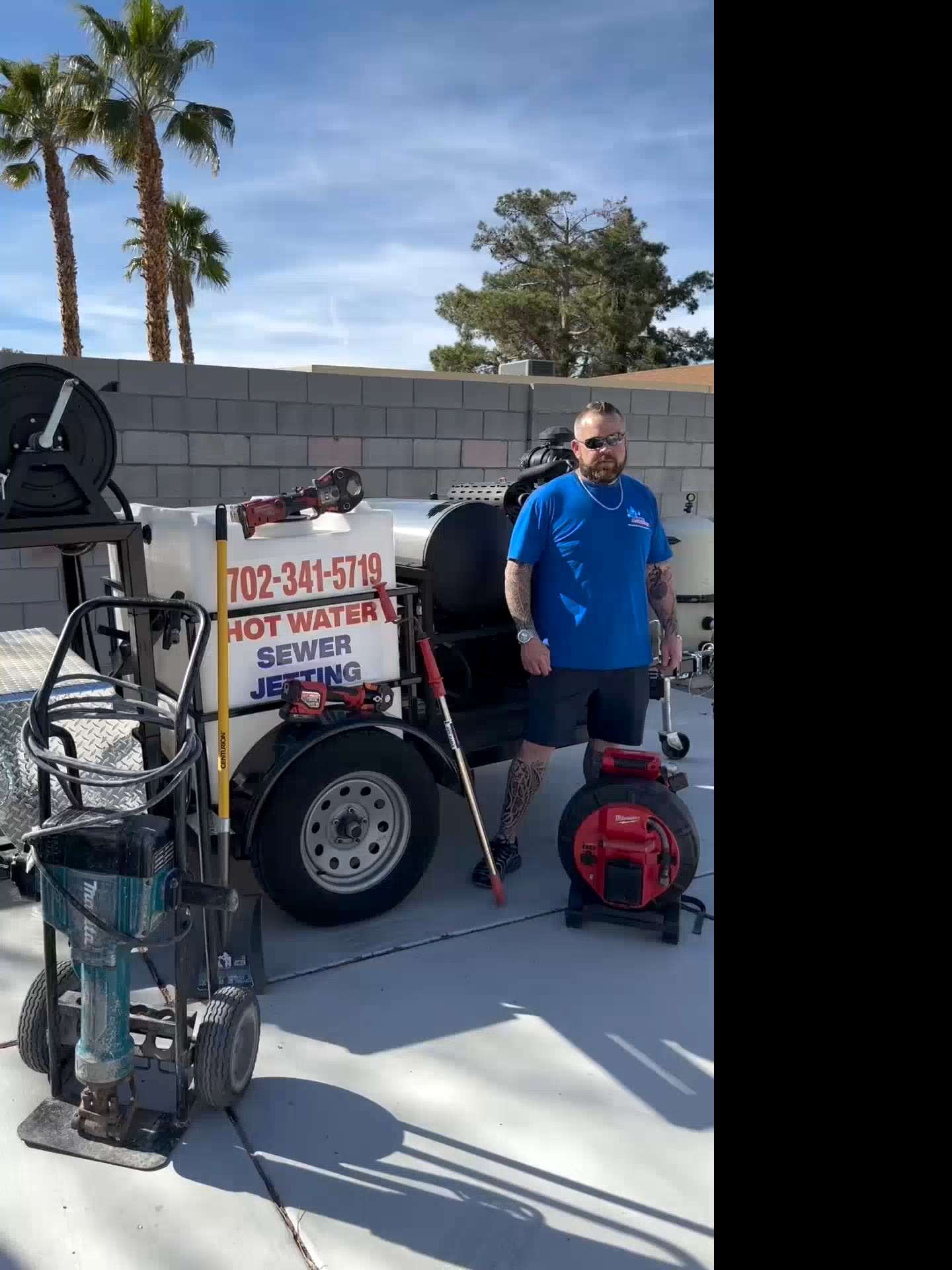 A crew member beside MC Plumbing's hot-water sewer-jetting trailer under palm trees.