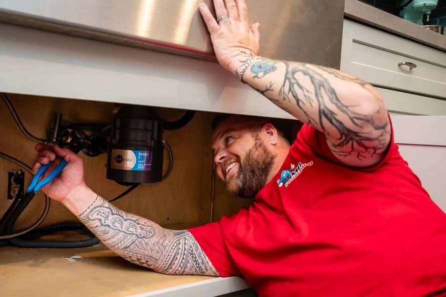 An MC Plumbing plumber working under a kitchen sink with blue pliers.